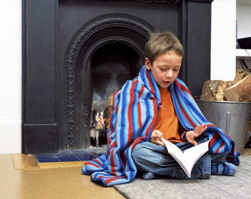 Boy in blanket on floor reading book by fireplace